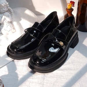 May include: A pair of glossy black loafers with a square toe and a chunky sole. The shoes feature a strap across the top with a gold-colored button detail. The shoes are on a white surface.