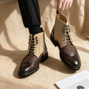 Op de afbeelding: Tweekleurige leren laarzen met een wingtip-ontwerp. De laarzen hebben een bruine neus en hiel, een beige bovenwerk en olijfgroene veters. De laarzen hebben een dikke zwarte zool. Getoond in een studio.