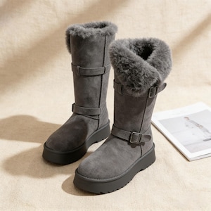 Puede incluir: Un par de botas de invierno grises con ribete de piel sintética. Las botas tienen una altura media de pantorrilla, una suela plana y dos correas ajustables con hebilla. Las botas están hechas de un material suave similar al ante.