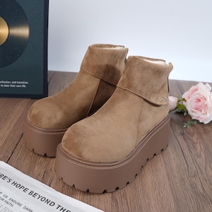 May include: A pair of tan suede platform boots with a folded-over cuff. The boots have a thick, brown sole and a soft, textured appearance. The background includes a dark green box with text and a pink rose.