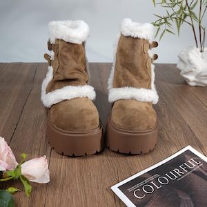 Puede incluir: Botas de plataforma marrones con ribete de piel sintética blanca y hebillas decorativas. Las botas tienen una suela gruesa marrón y se exhiben sobre una superficie de madera. Las botas miden unos 10 cm de alto.