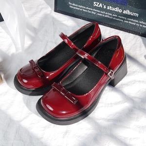 Peut inclure: Une paire de chaussures Mary Jane bordeaux vernies, avec un nœud sur le bout et une bride de cheville. Les chaussures ont un talon et une semelle noirs épais. Le texte en arrière-plan indique "SZA's studio album".
