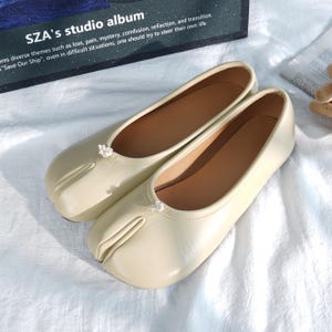 Peut inclure: Une paire de ballerines à bout fendu de couleur crème. Les chaussures ont un bout arrondi avec une fente pour le gros orteil et un petit nœud décoratif sur le dessus. Les chaussures sont sur une surface blanche. Le texte "SZA's studio album" est visible en arrière-plan.