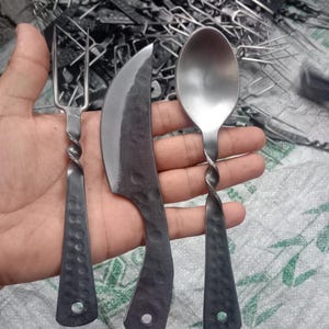 May include: Hand-forged metal cutlery set including a fork, knife, and spoon. The handles have a twisted design and a hammered texture. The knife has a curved blade. The set is made of dark metal.