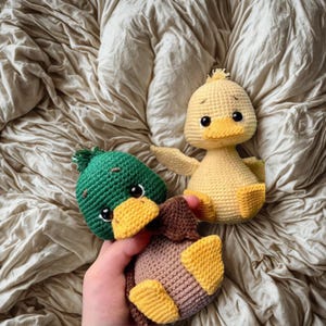 May include: Three handmade crochet ducklings in the image. One is yellow, one is green and brown, and the third is brown and yellow. The ducklings have black eyes and yellow beaks. The ducklings are sitting on a wrinkled, beige fabric.