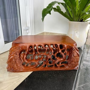 May include: A rectangular, reddish-brown wooden display stand with intricate carved openwork on the sides. The top surface is smooth, while the sides feature a detailed, organic design. The stand is likely used for decorative items.