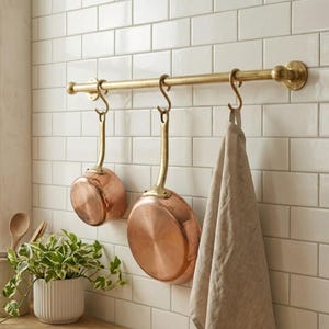 Puede incluir: Un estante de latón con dos ollas de cobre y un paño de cocina beige colgando. El estante está montado en una pared de azulejos blancos. Una pequeña planta en maceta se encuentra sobre una superficie de madera.