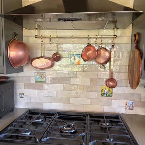 Może przedstawiać: Scena kuchenna z czarną kuchenką gazową, okapem i mosiężnym stojakiem na garnki. Miedziane garnki i patelnie o różnych rozmiarach wiszą na stojaku. Widoczne są również dekoracyjne płytki i drewniana deska do krojenia.