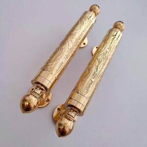 Puede incluir: Dos manijas de puerta doradas y ornamentadas. Cada manija presenta intrincados patrones grabados a lo largo de su cuerpo cilíndrico y está montada sobre una base circular. Las manijas tienen un extremo redondeado y decorativo.