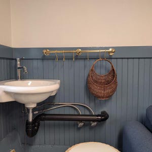 Puede incluir: Un cuarto de baño con un lavabo de porcelana blanca y un grifo cromado. Una barra de color latón con ganchos cuelga encima, y una cesta de mimbre está suspendida de uno de los ganchos. La pared está pintada de azul.