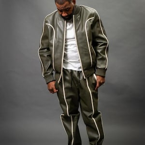 K&ouml;nnte beinhalten: Olivgr&uuml;ne Lederjacke und passende Hose mit wei&szlig;em Besatz. Die Jacke hat einen Rei&szlig;verschluss und gerippte B&uuml;ndchen. Die Hose hat einen Kordelzug in der Taille. Das Outfit wird mit wei&szlig;en Turnschuhen kombiniert.