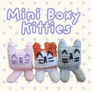 May include: Three small crocheted cat figures labeled Mini Boxy Kitties. One is light pink, one is white and orange, and one is light gray. Each has two pointed ears, two dangling legs, and embroidered facial features including eyes and whiskers.