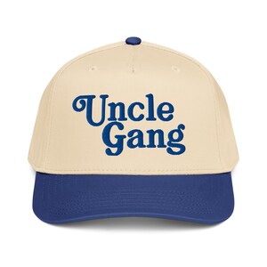 Puede incluir: Gorra de béisbol con corona color crema y visera azul marino. La gorra presenta el texto "Uncle Gang" en una fuente azul marino en negrita en la parte delantera. La gorra tiene una visera curva y un botón en la parte superior.