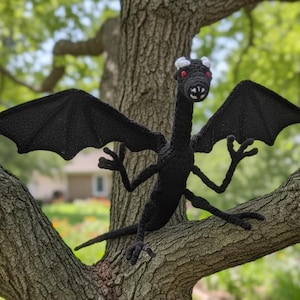 May include: A black crocheted dragon with large wings, perched on a tree branch. The dragon has white horns and red eyes. The background is blurred, showing a house and greenery.