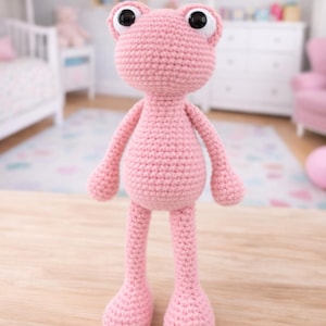May include: A handmade, pink crochet frog doll with large black eyes. The frog has a rounded body, long legs, and small arms. The doll is standing on a wooden surface, with a blurred background of a room.