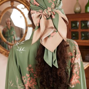 May include: A green silk scarf with a floral pattern is tied around the head, with a contrasting tan panel. The scarf is paired with a green floral robe. The image is taken from behind, showcasing the back of the head and robe.