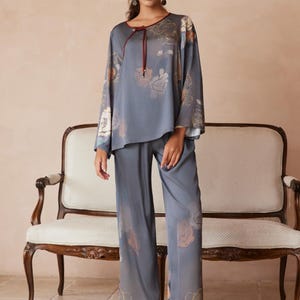 May include: A two-piece pajama set in a muted blue-gray hue, adorned with floral patterns in shades of peach and gold. The top features a round neckline with a burgundy tie, and the wide-leg pants complete the ensemble. The model is wearing pink floral slippers.