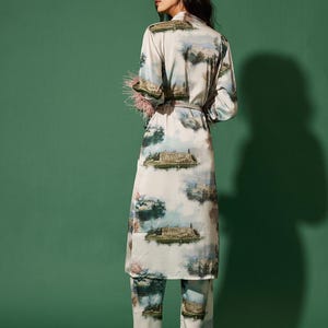 May include: A light-coloured pajama set featuring a long-sleeved robe and matching trousers. The set is adorned with a scenic landscape print in shades of blue, green, and beige. The robe has a belt and is trimmed with pink feathers.