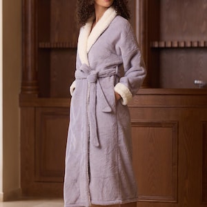 Peut inclure: Un peignoir lilas avec un col et des poignets blancs et duveteux. Le peignoir présente un motif texturé et une ceinture assortie. Le modèle porte le peignoir dans une pièce avec une armoire en bois. Le peignoir arrive au genou.