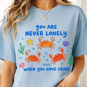 May include: A light blue short-sleeve t-shirt featuring a graphic print of three orange crabs, a yellow starfish, seashells, and green seaweed. The text reads: YOU ARE NEVER LONELY WHEN YOU HAVE CRABS. The design is colorful and hand-drawn in style.