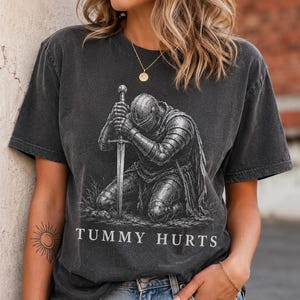May include: A charcoal gray short-sleeve t-shirt featuring a graphic of a knight in full armor kneeling and holding a sword. Below the illustration, the text "TUMMY HURTS" is printed in a white serif font. The shirt has a crew neckline.