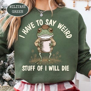 May include: Military green sweatshirt with the text "I HAVE TO SAY WEIRD STUFF OF I WILL DIE." The graphic features a frog wearing a hat and sweater, holding a cup. The sweatshirt is a comfortable, casual style.