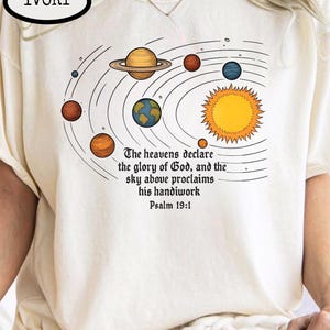 May include: Ivory-colored t-shirt featuring a solar system graphic with planets and a sun. The design includes the text "The heavens declare the glory of God, and the sky above proclaims his handiwork Psalm 19:1."
