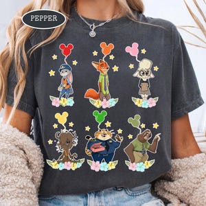 K&ouml;nnte beinhalten: Dunkelgraues T-Shirt mit Zeichentrickfiguren aus Zootopia, die jeweils einen Mickey Mouse-f&ouml;rmigen Ballon halten. Die Figuren sind vor einem Hintergrund aus Sternen und Blumen platziert. Das Wort "PEPPER" ist oben links aufgedruckt.