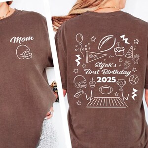 May include: Brown t-shirt with the word "Mom" and a football helmet graphic on the front. The back of the shirt features a football-themed design with the text "Elijah's First Birthday 2025".