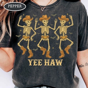 May include: Dark gray t-shirt featuring a graphic of three dancing skeletons wearing cowboy hats and boots. The phrase "YEE HAW" is printed below the skeletons. The shirt has a vintage, distressed look.