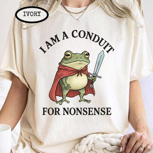 May include: Ivory-colored t-shirt featuring a cartoon frog in a red cape holding a sword. The text on the shirt reads "I AM A CONDUIT FOR NONSENSE". The shirt has short sleeves and a crew neck.