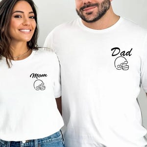 May include: White t-shirts with black text and a football helmet graphic. One shirt says "Mom" and the other says "Dad". Both shirts are paired with blue jeans. The shirts are a matching set.