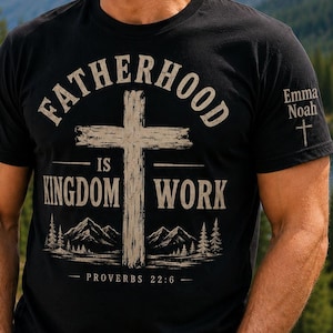 May include: A black short-sleeved t-shirt featuring a graphic with a large cross, mountains, and pine trees. The text reads "FATHERHOOD IS KINGDOM WORK" and "PROVERBS 22:6". The left sleeve displays the names "Emma" and "Noah" above a small cross.