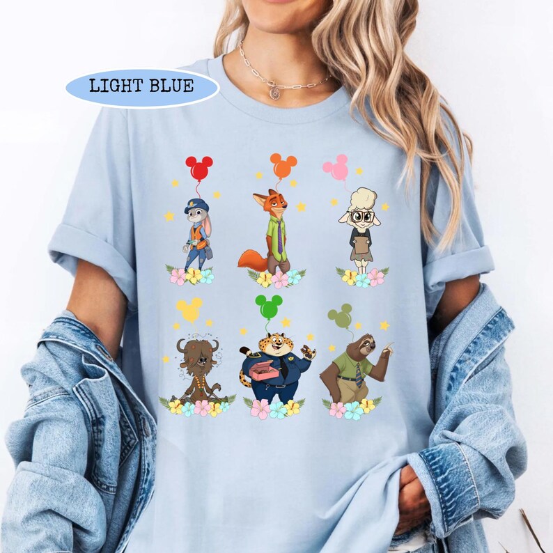 K&ouml;nnte beinhalten: Hellblaues T-Shirt mit Zeichentrickfiguren aus Zootopia. Das Shirt hat einen Rundhalsausschnitt und kurze &Auml;rmel. Die Charaktere sind von Blumen und Luftballons umgeben. Der Text "LIGHT BLUE" befindet sich in einem hellblauen Rechteck oben.