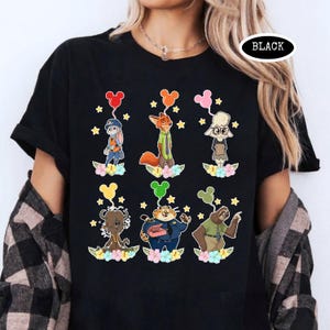 K&ouml;nnte beinhalten: Schwarzes T-Shirt mit Zeichentrickfiguren aus Zootopia, die jeweils einen Mickey-Mouse-f&ouml;rmigen Ballon halten. Die Figuren sind vor einem Hintergrund aus Blumen und Sternen platziert. Das Wort "BLACK" ist auf dem Shirt aufgedruckt.