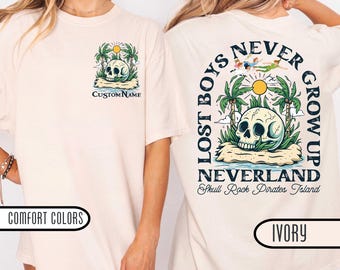 May include: Ivory-colored t-shirt featuring a skull on a tropical island with palm trees and the text "Lost Boys Never Grow Up" on the back. The front has a similar design with "Custom Name" text. The shirt is part of the "Comfort Colors" collection.