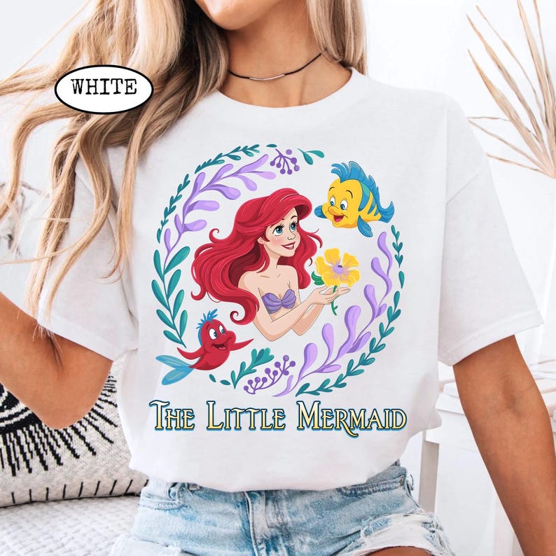 Puede incluir: Camiseta blanca con un gr&aacute;fico colorido de Ariel de La Sirenita. El dise&ntilde;o incluye a Ariel, Flounder, Sebasti&aacute;n y una flor amarilla, rodeados por una corona floral. Las palabras "The Little Mermaid" est&aacute;n impresas debajo.