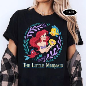 Puede incluir: Camiseta negra con un gr&aacute;fico colorido de Ariel de La Sirenita, rodeada por una corona floral. El dise&ntilde;o incluye a Flounder, Sebasti&aacute;n y el texto "The Little Mermaid". Una etiqueta ovalada negra est&aacute; en la esquina superior derecha.