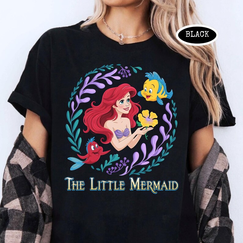 Puede incluir: Camiseta negra con un gr&aacute;fico colorido de Ariel de La Sirenita, rodeada por una corona floral. El dise&ntilde;o incluye a Flounder, Sebasti&aacute;n y el texto "The Little Mermaid". Una etiqueta ovalada negra est&aacute; en la esquina superior derecha.