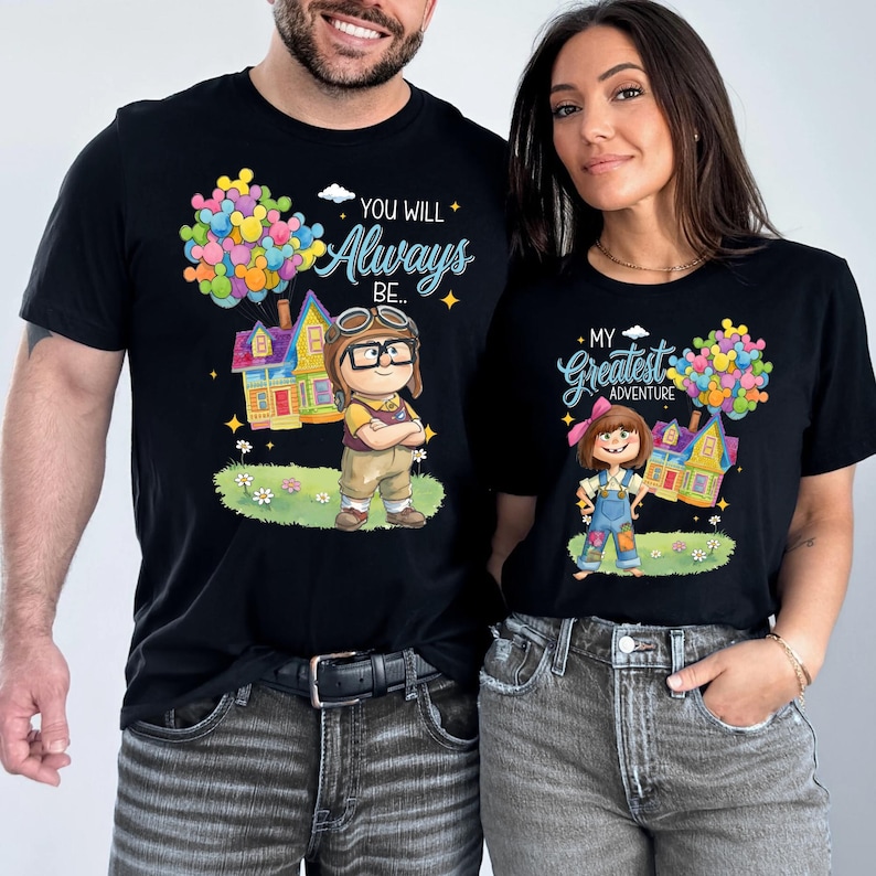 K&ouml;nnte beinhalten: Schwarze T-Shirts mit passenden Grafiken. Ein T-Shirt zeigt den Text "You Will Always Be..." mit einem Cartoon-Bild eines Hauses und einer Figur. Das andere Shirt sagt "My Greatest Adventure" mit einem &auml;hnlichen Design.
