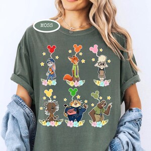 K&ouml;nnte beinhalten: Gr&uuml;nes T-Shirt mit Zeichentrickfiguren aus Zootopia, die jeweils einen Ballon halten. Die Charaktere sind in zwei Reihen angeordnet, mit floralen und Sternen-Akzenten. Das Wort "MOSS" ist auf dem Shirt aufgedruckt.
