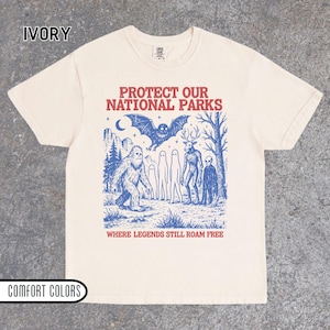 May include: An ivory short-sleeve t-shirt featuring a blue graphic print of a bat, a sasquatch, ghosts, a deer-headed creature, and an alien. The text reads PROTECT OUR NATIONAL PARKS and WHERE LEGENDS STILL ROAM FREE in red lettering.