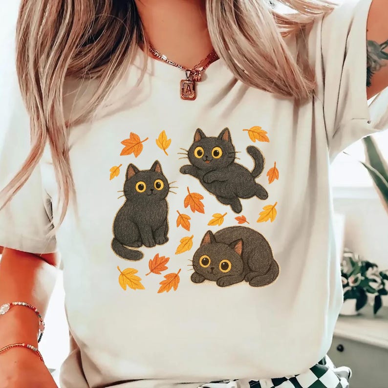 May include: A cream-colored t-shirt featuring a playful design of three black cats with yellow eyes and autumn leaves. One cat is sitting, another is leaping, and the third is curled up. The leaves are in shades of yellow and orange, creating a fall theme.