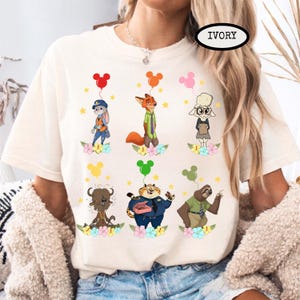 K&ouml;nnte beinhalten: Elfenbeinfarbenes T-Shirt mit Cartoon-Charakteren aus Zootopia, die jeweils einen Ballon halten. Die Charaktere sind von Blumen und Sternen umgeben. Das Wort "IVORY" ist oben rechts auf dem Shirt aufgedruckt.