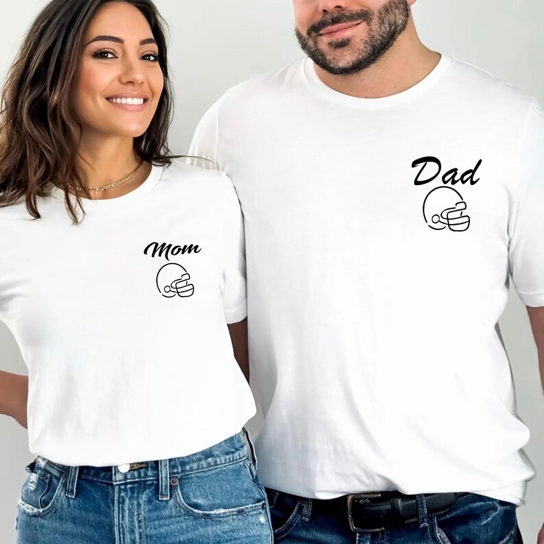 May include: White t-shirts with black text and a football helmet graphic. One shirt says "Mom" and the other says "Dad". Both shirts are paired with blue jeans. The shirts are a matching set.