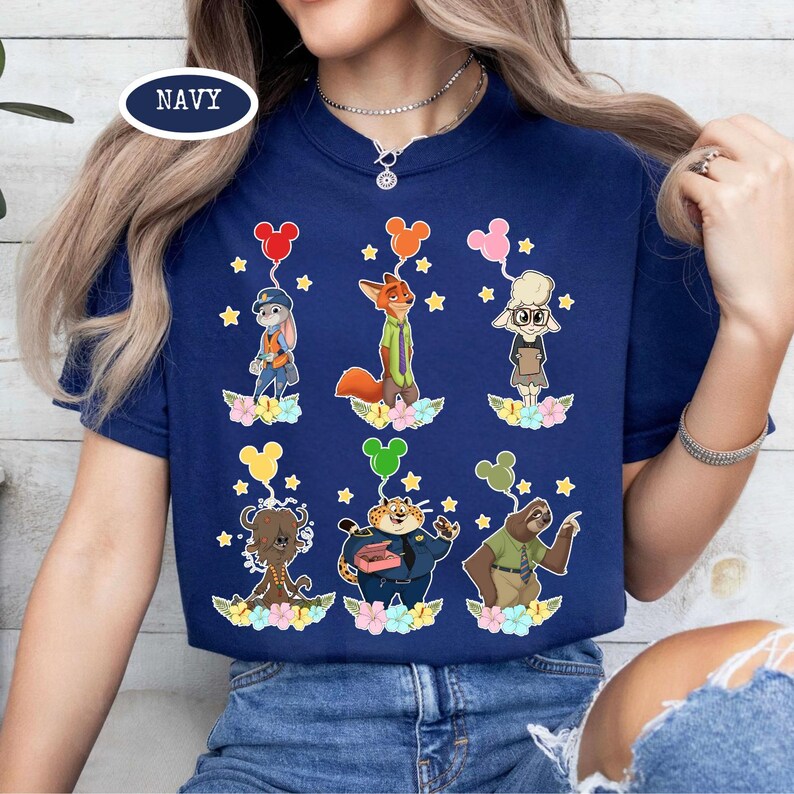 K&ouml;nnte beinhalten: Marineblaues T-Shirt mit Zeichentrickfiguren aus Zootopia, jede mit einem Mickey Mouse-Ballon. Das Design umfasst Judy Hopps, Nick Wilde und andere Charaktere, mit floralen und Sternendetails. Das Wort "NAVY" ist oben links aufgedruckt.