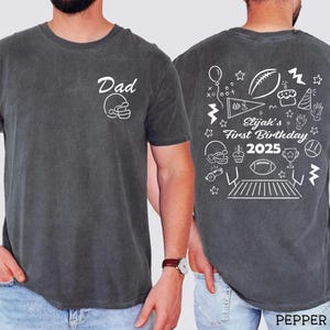 May include: Dark gray t-shirt with white graphic text and illustrations. The front features "Dad" and a football helmet. The back displays "Elijah's First Birthday 2025" with football-themed drawings, including a football, cake, and trophy.