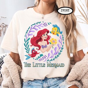 Puede incluir: Camiseta de color marfil con una ilustraci&oacute;n colorida de Ariel de La Sirenita. El dise&ntilde;o incluye a Ariel, Flounder, Sebasti&aacute;n y una flor amarilla, rodeados por una corona floral. El texto "The Little Mermaid" est&aacute; impreso debajo.