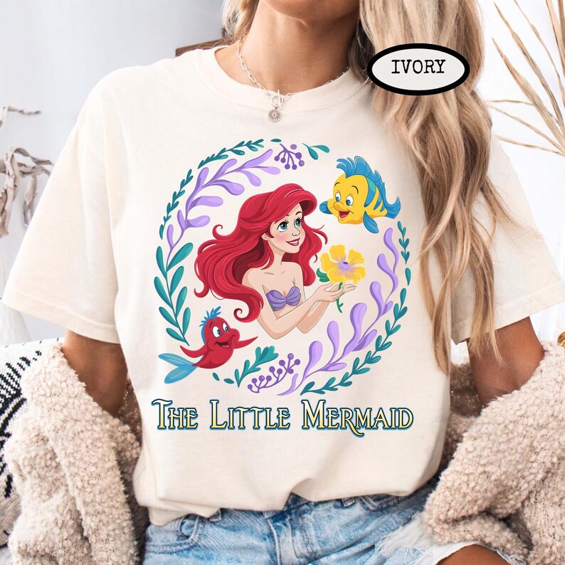 Puede incluir: Camiseta de color marfil con una ilustraci&oacute;n colorida de Ariel de La Sirenita. El dise&ntilde;o incluye a Ariel, Flounder, Sebasti&aacute;n y una flor amarilla, rodeados por una corona floral. El texto "The Little Mermaid" est&aacute; impreso debajo.