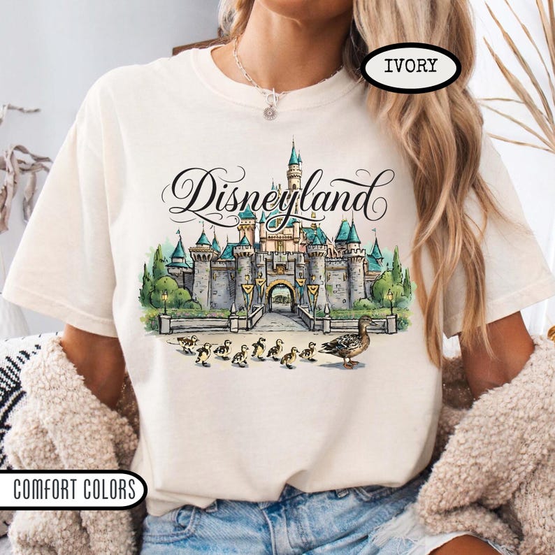May include: Ivory-colored t-shirt featuring a Disneyland castle graphic with the word "Disneyland" in script. The graphic includes ducks and ducklings. The shirt is labeled "Comfort Colors".